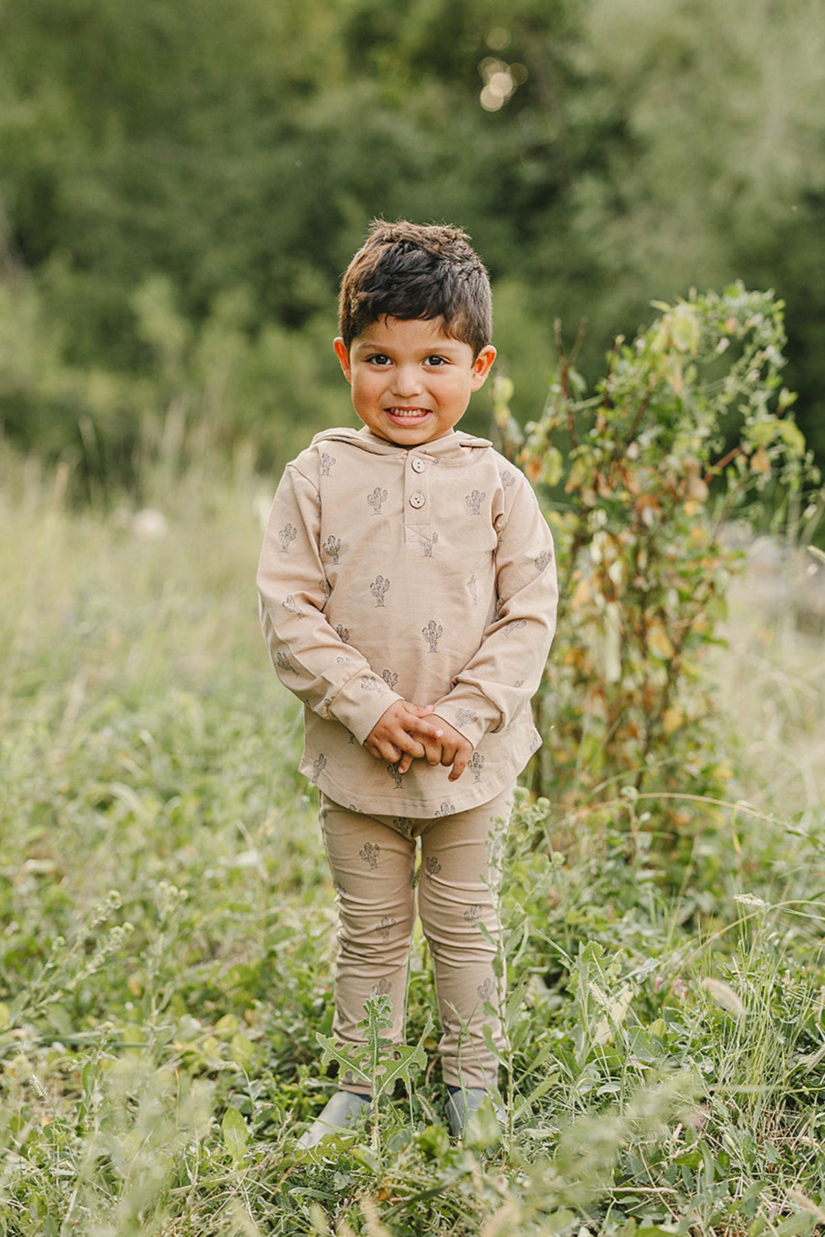 A child wearing a beige cactus-patterned long-sleeve hooded set stands smiling in a grassy outdoor area.