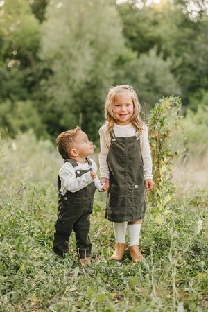 Two young children wearing green corduroy overalls stand in a lush, green outdoor setting, smiling and holding hands.