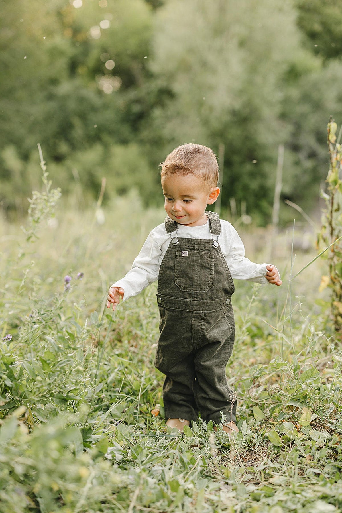 A toddler in green corduroy overalls and white shirt explores a lush, grassy field outdoors.