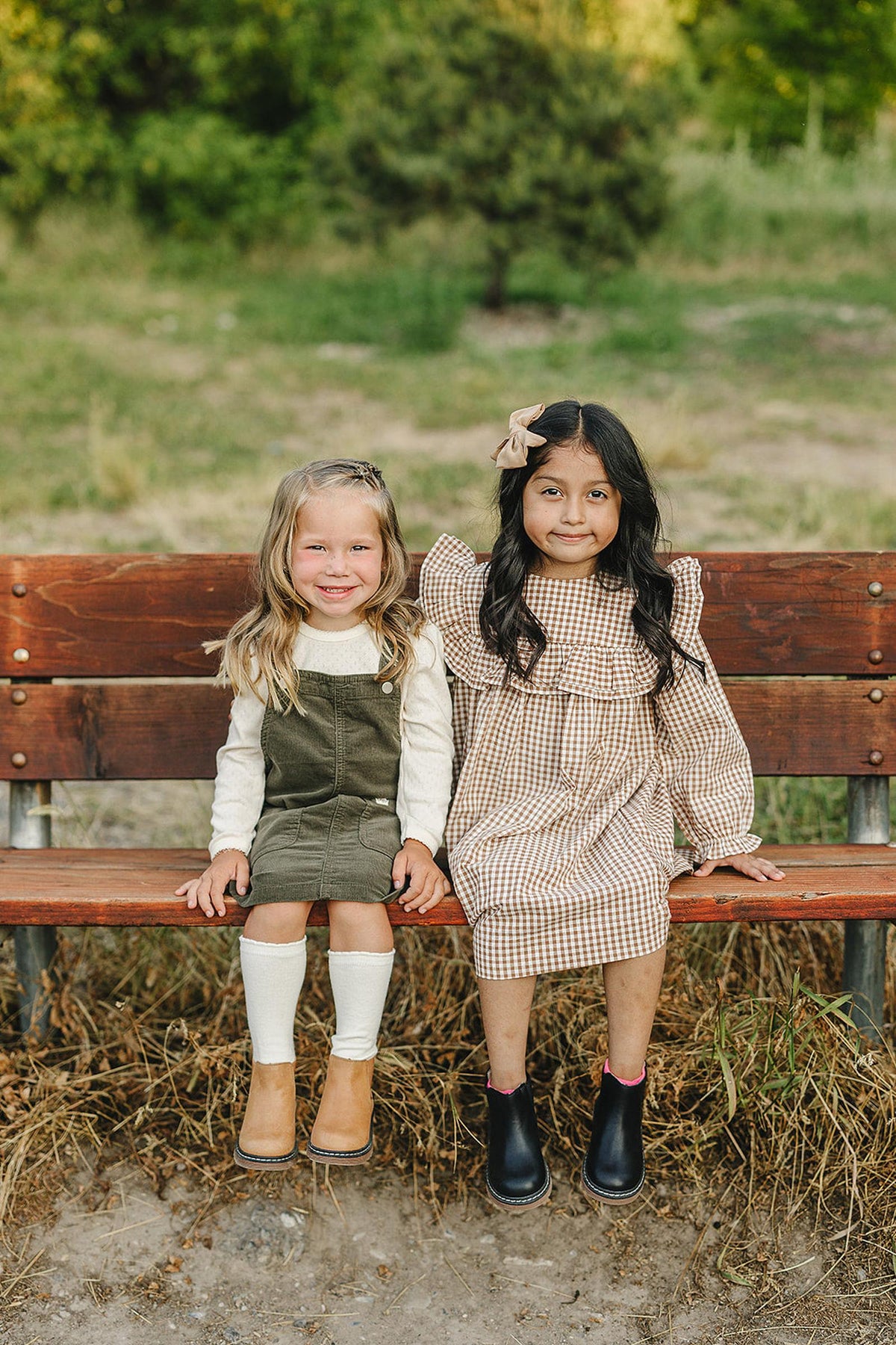 Two children sit on a wooden bench; one wears a green corduroy dress, the other a plaid dress.
