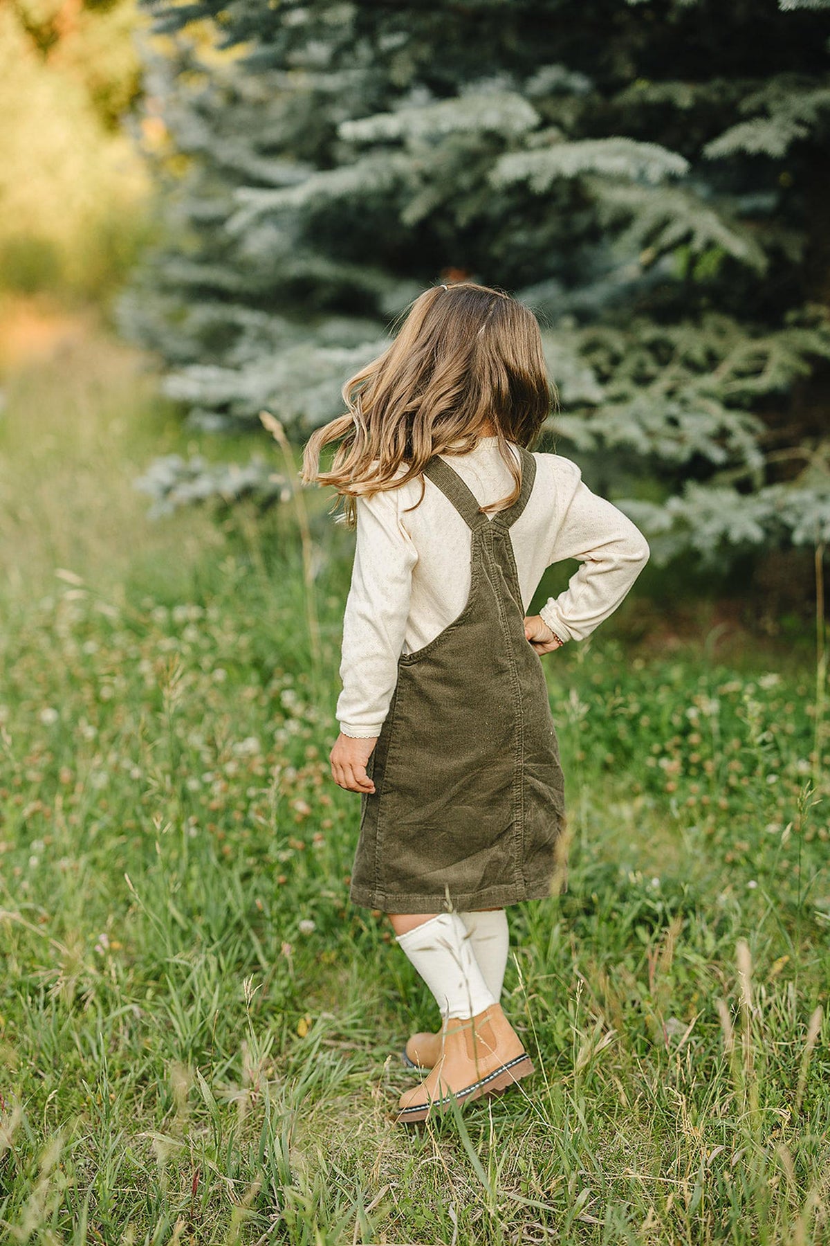 A child in a green corduroy dress stands outdoors, wearing brown boots and white tights, surrounded by greenery.