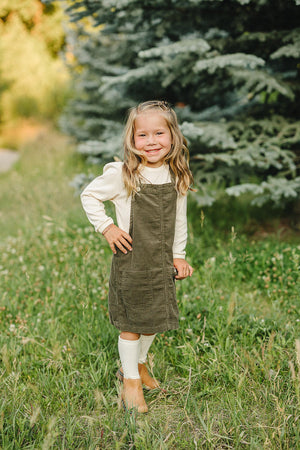 A young girl stands in a grassy area wearing a green corduroy dress and cream shirt, smiling brightly.