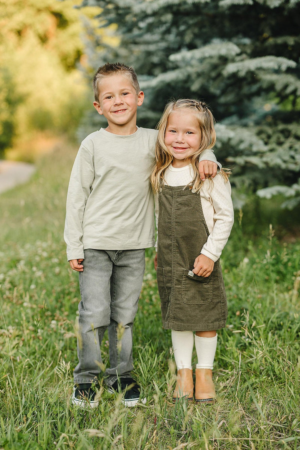 Two smiling children stand in a grassy field, one wearing a green corduroy dress, with trees in the background.