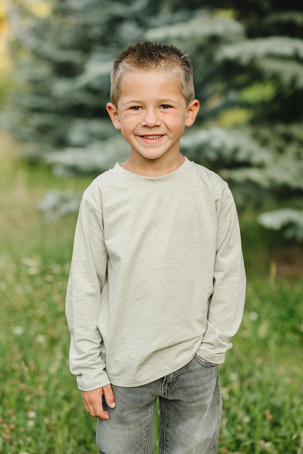 A smiling child wearing a sage long sleeve cotton T-shirt stands outdoors with greenery and trees in the background.