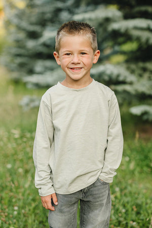 A smiling child wearing a sage long sleeve cotton T-shirt stands outdoors with greenery and trees in the background.