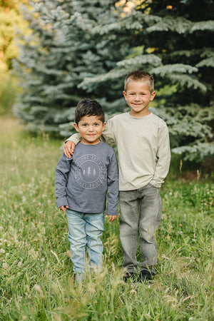 Two young boys stand in a grassy field, wearing long-sleeve shirts, one grey and one light sage.