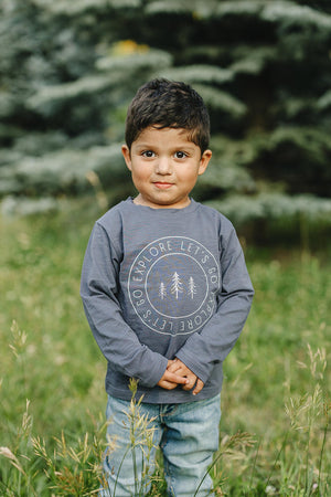 A child wearing a navy Lets Go Explore long sleeve t-shirt stands in a grassy outdoor setting.