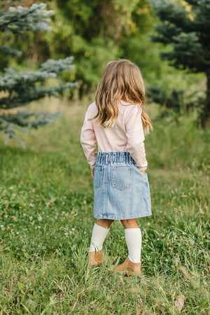A child in a pink long sleeve shirt and denim skirt stands in a grassy outdoor setting.