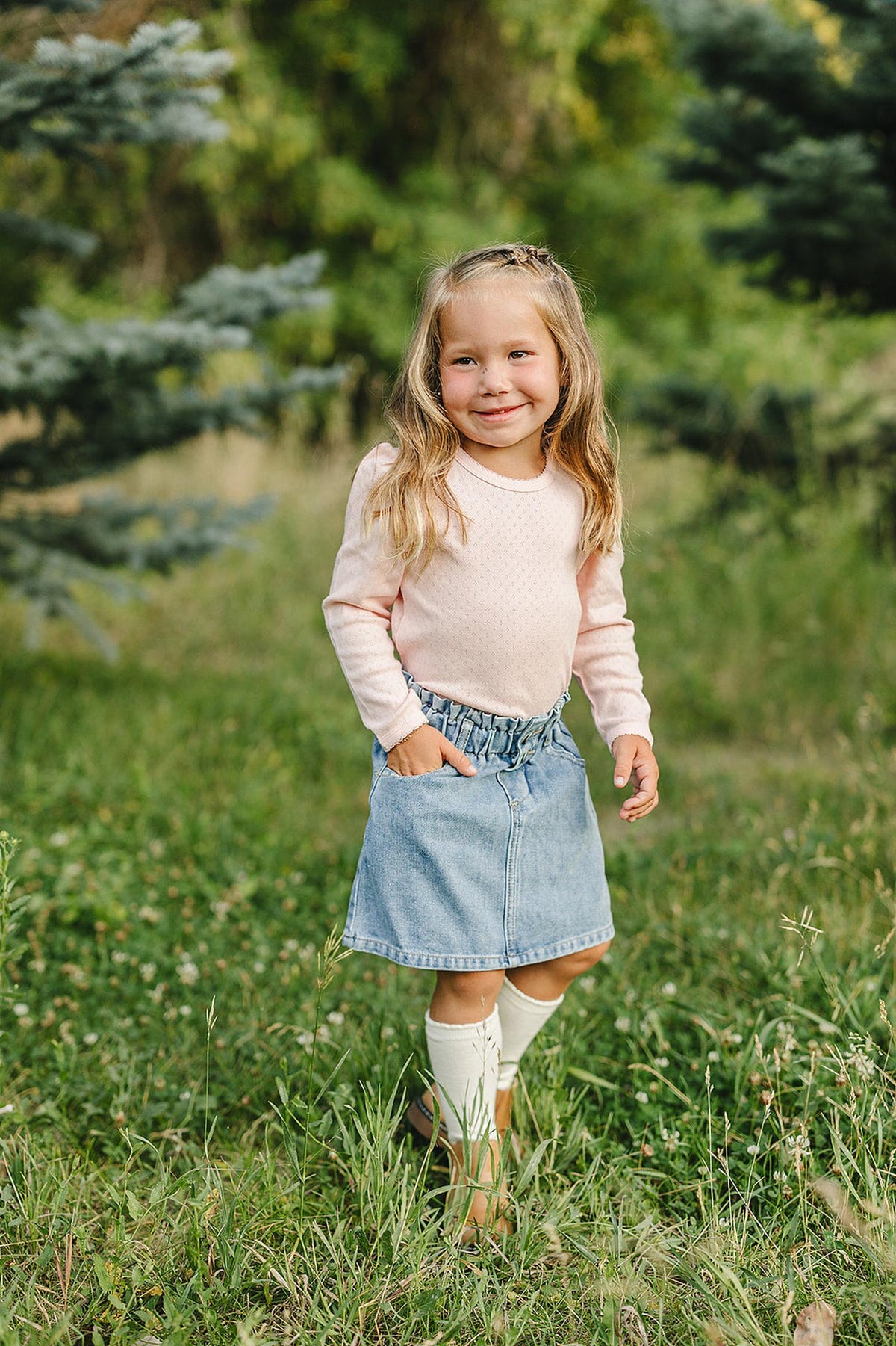A young girl in a pink pointelle long sleeve shirt and denim skirt stands in a grassy field.