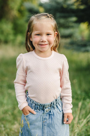 A child smiles while wearing a pink pointelle long sleeve shirt and denim skirt, standing in a grassy field.