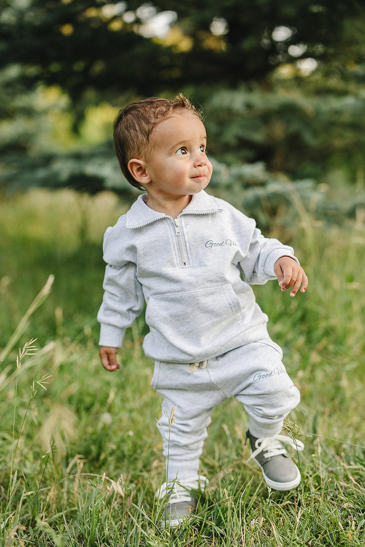 A toddler wearing a grey quarter-zip sweatshirt and pants set with Good Vibes text, standing in a grassy field.