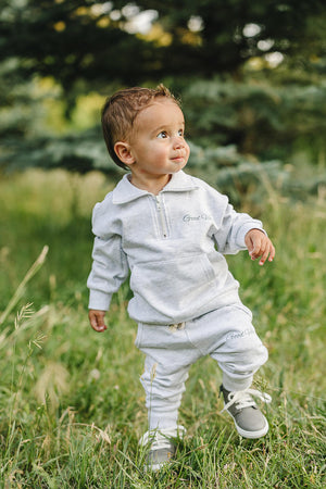 A toddler wearing a grey quarter-zip sweatshirt and pants set with Good Vibes text, standing in a grassy field.