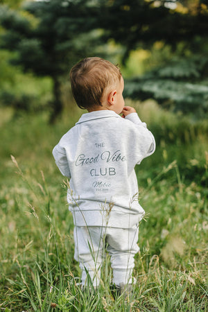 Toddler in a grey sweatshirt with Good Vibes Club text, standing in a grassy outdoor setting.