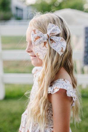 A young girl with long, wavy hair wears a large, handcrafted bow featuring a western pony print. The bow is made from cotton fabric and adds charm to her outfit.