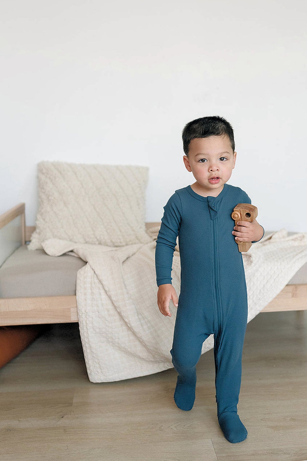A toddler wearing a navy ribbed zipper footed pajama holds a wooden toy in a cozy room.