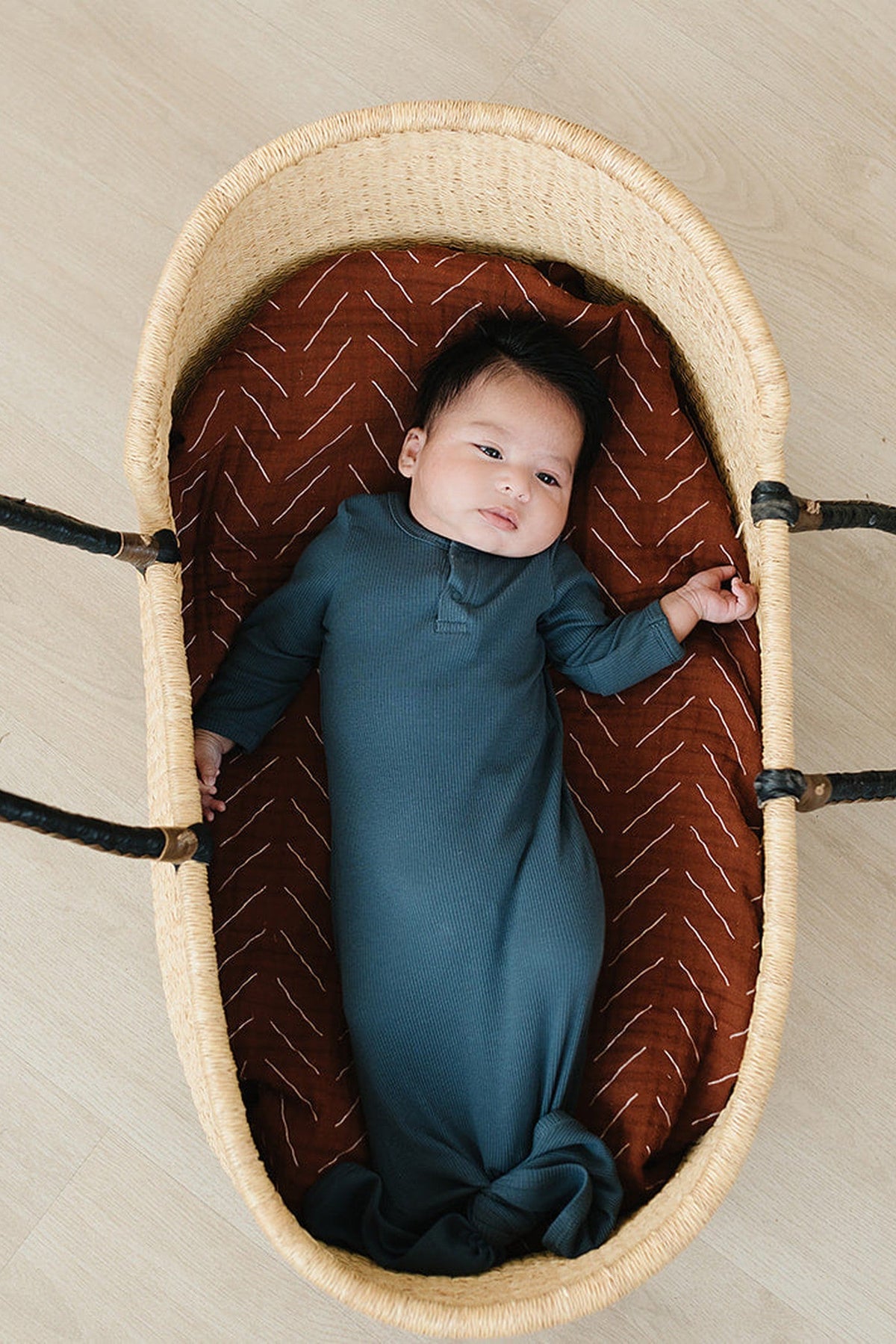 A baby in a navy ribbed gown lies in a woven basket with a brown patterned lining.