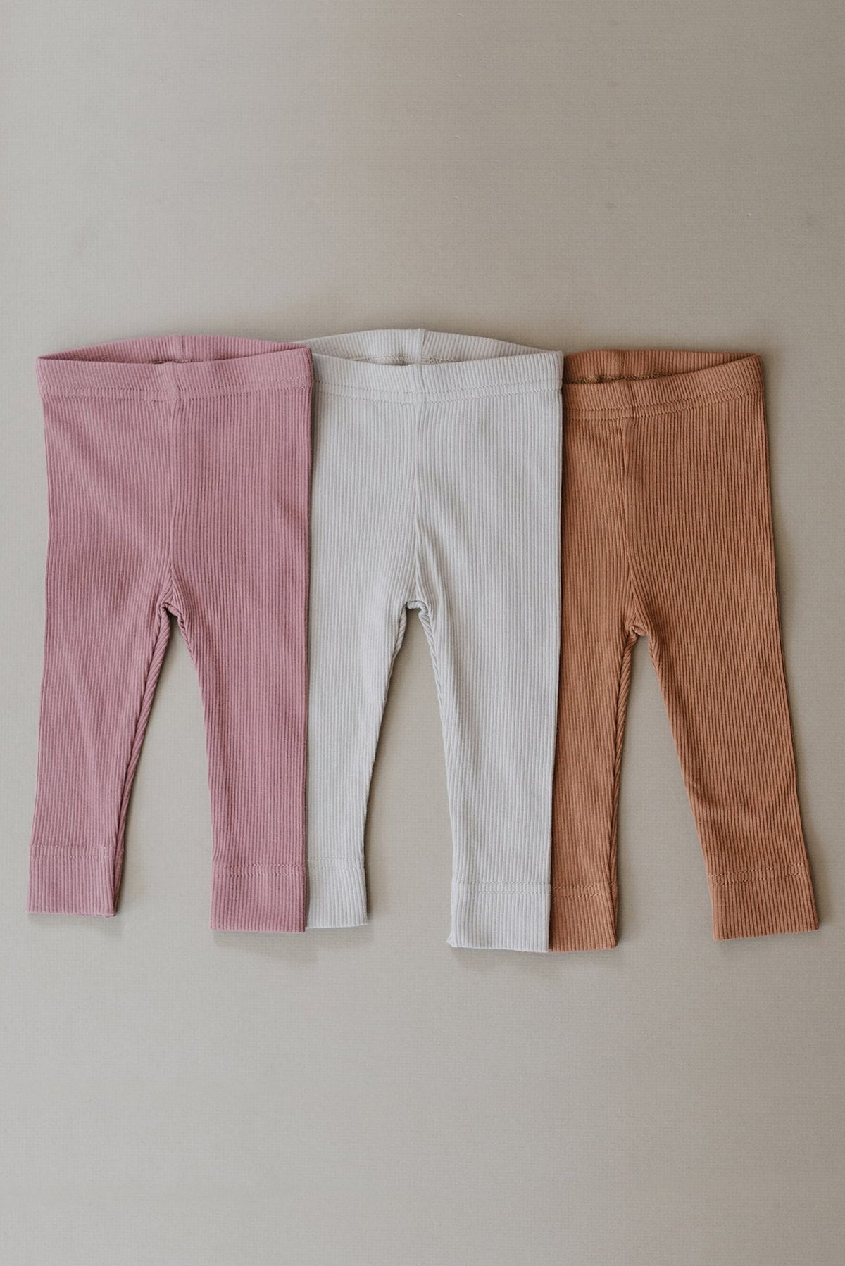 Three pairs of ribbed leggings in pink, white, and brown colors are displayed side by side on a plain background.