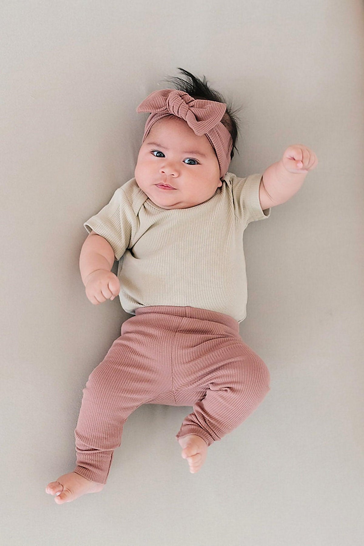 Baby in dusty rose leggings and headband with a cream shirt, lying on a neutral background.