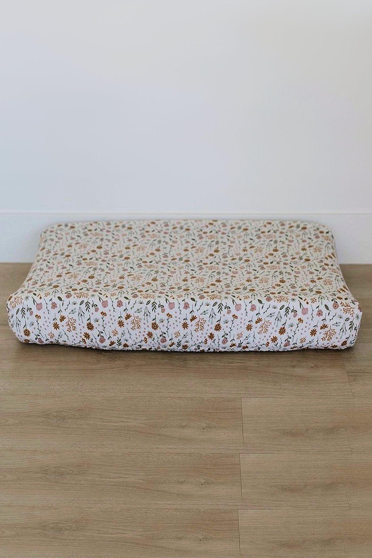 Floral muslin changing pad cover with a delicate pattern sits on a wooden floor in a bright room.