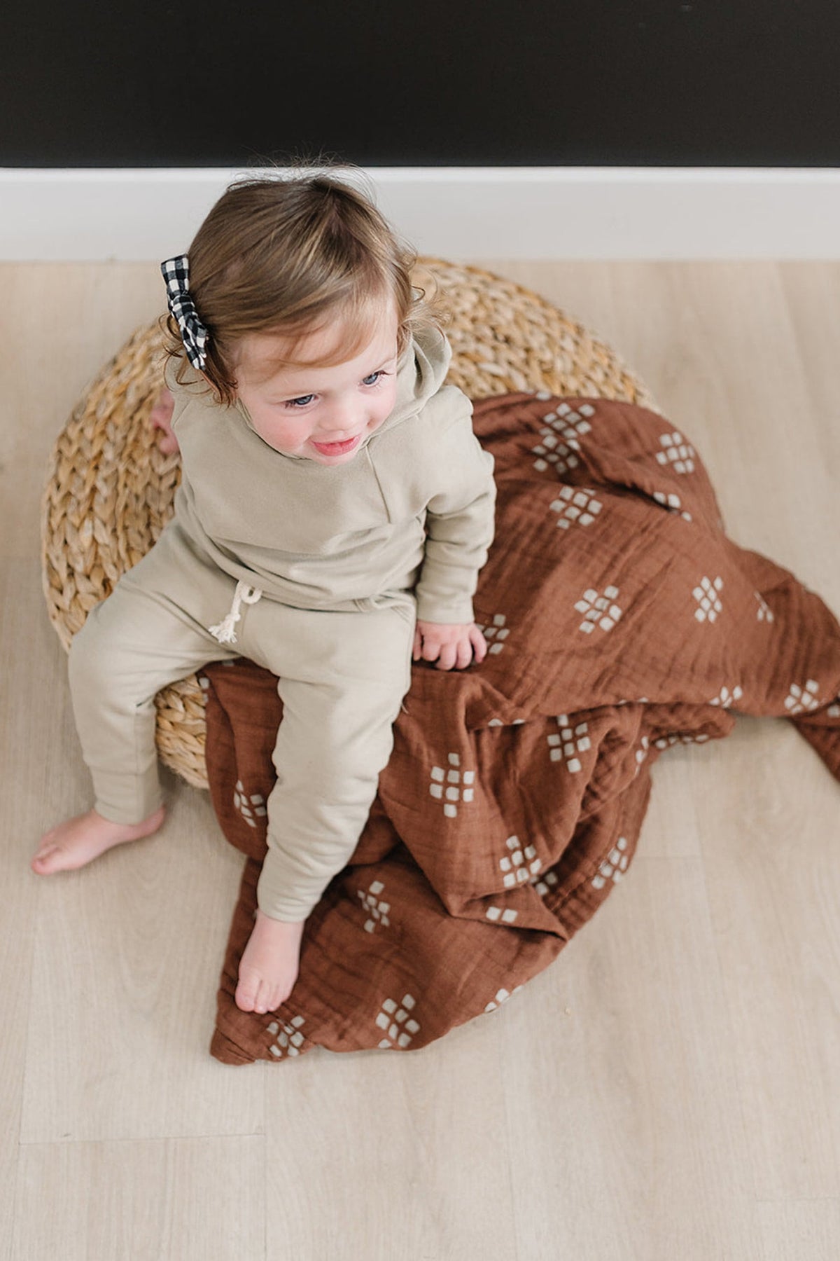 A child in a beige outfit sits on a woven basket with a brown patterned muslin quilt.