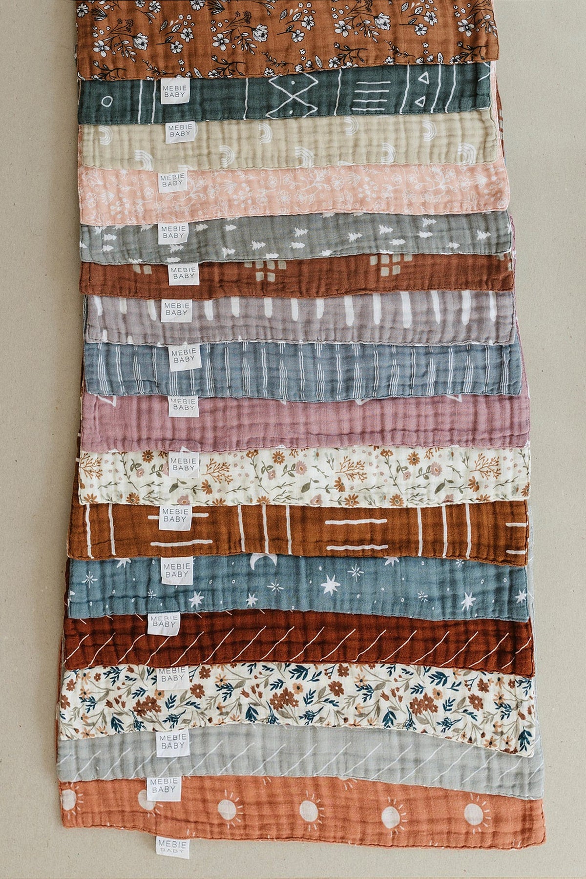 A collection of Mebie Baby muslin burp cloths in various patterns and colors, neatly stacked on a surface.
