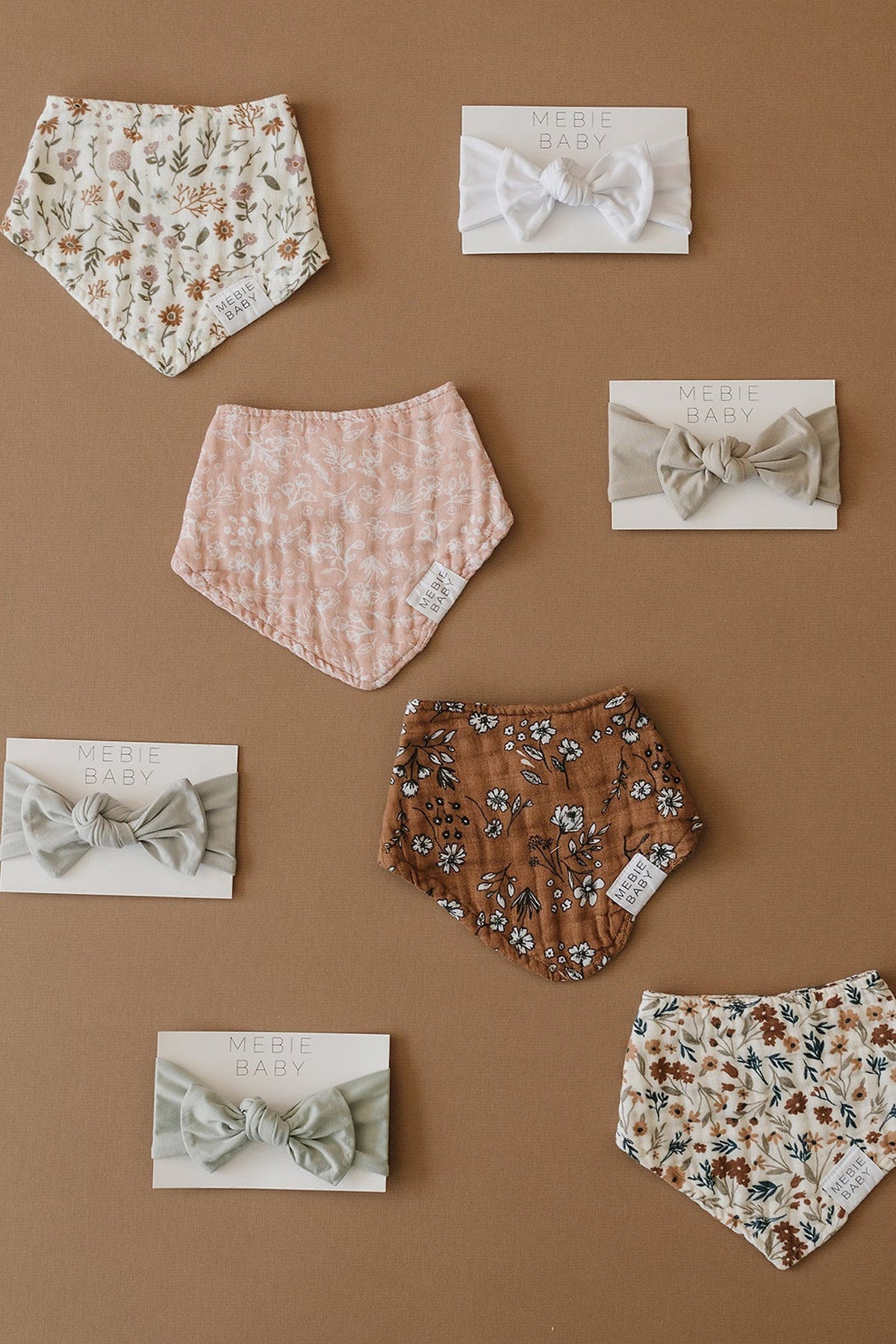 Various Mebie Baby muslin bibs and bows displayed on a brown background, featuring floral patterns and earthy colors.