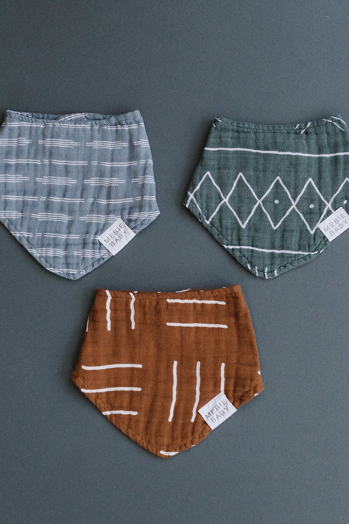 Three Mebie Baby muslin bibs in gray, green, and mustard with geometric patterns on a dark background.