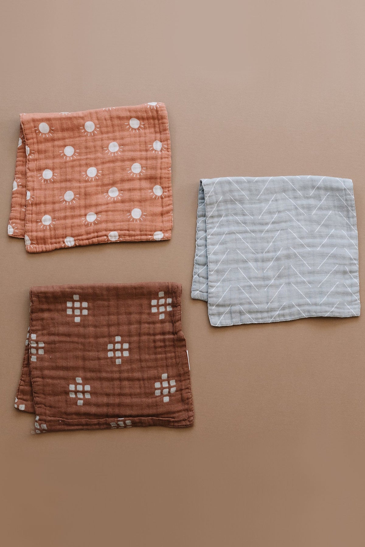Three muslin burp cloths in brown, chestnut, and gray with various patterns are displayed on a beige surface.