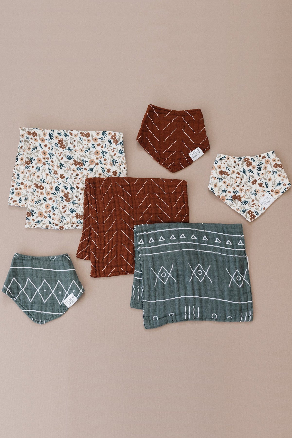 Assorted baby bibs and cloths in rust, green, and floral patterns laid on a beige surface.