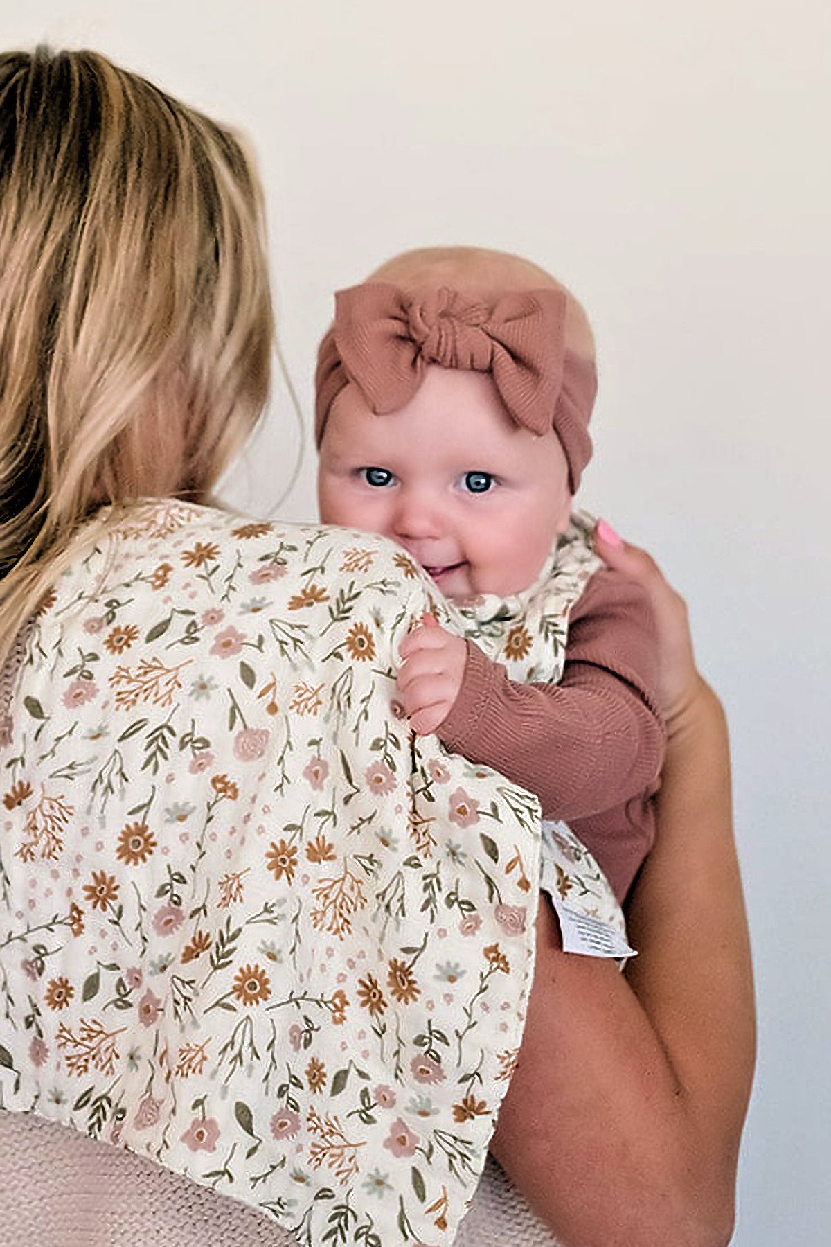 A baby wearing a dusty rose ribbed head wrap is held by a woman, with a floral-patterned cloth nearby.
