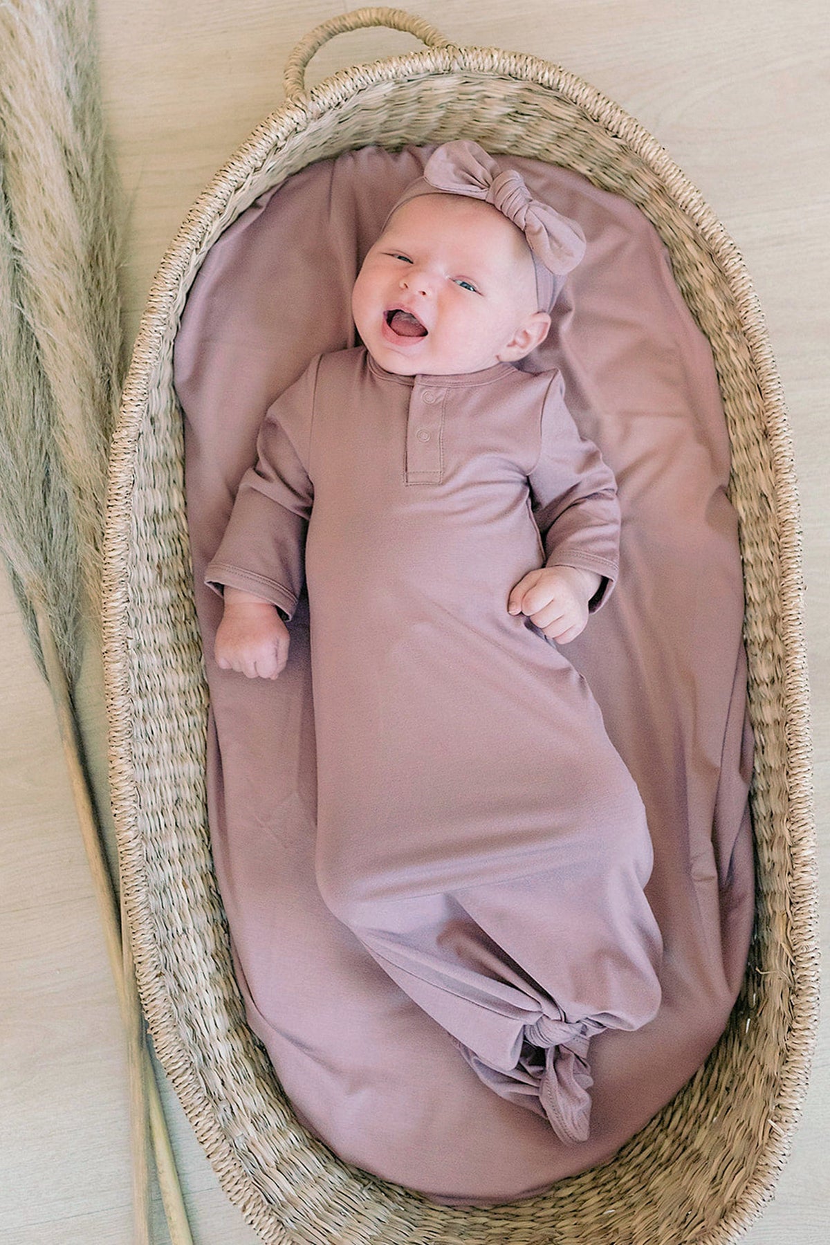 A baby in a dusty rose knot gown and headband lies in a woven basket, smiling contentedly.