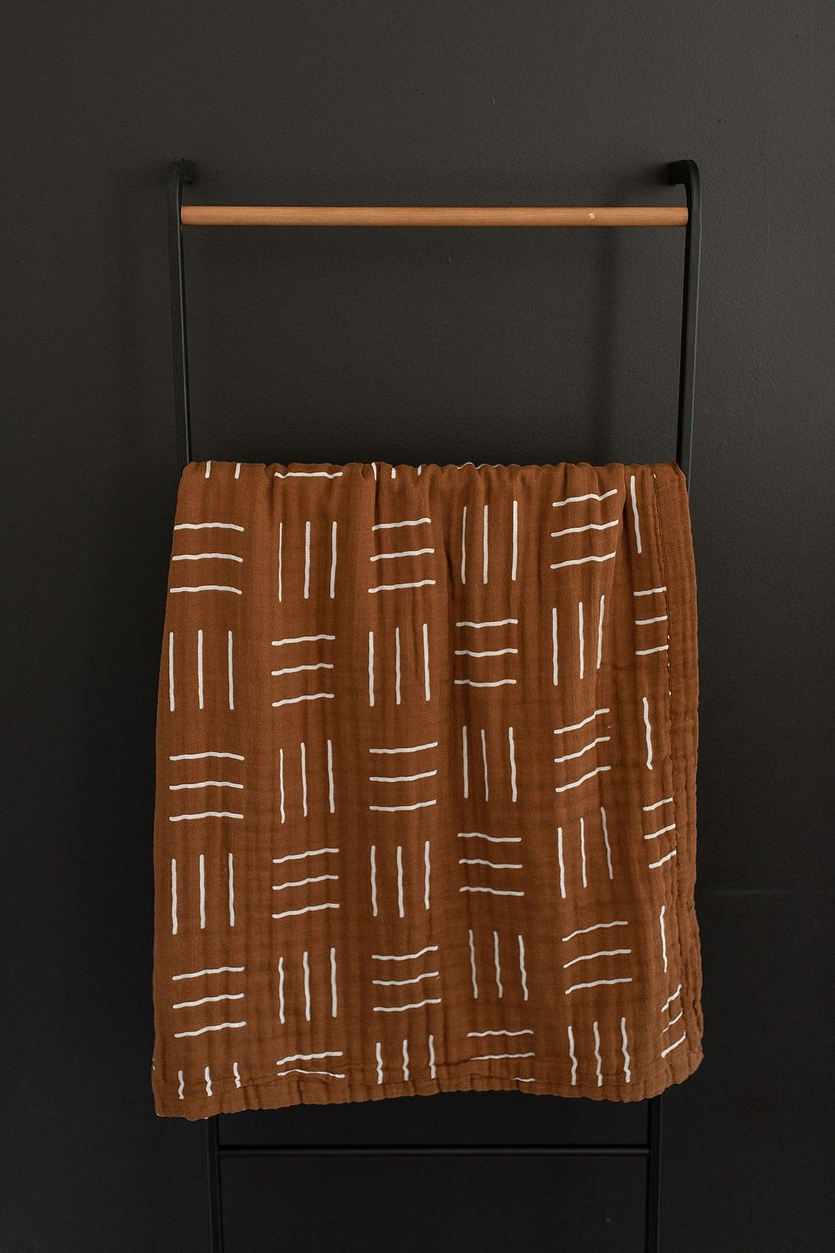 A mustard-colored muslin quilt with white mudcloth patterns hangs on a black and wooden rack.