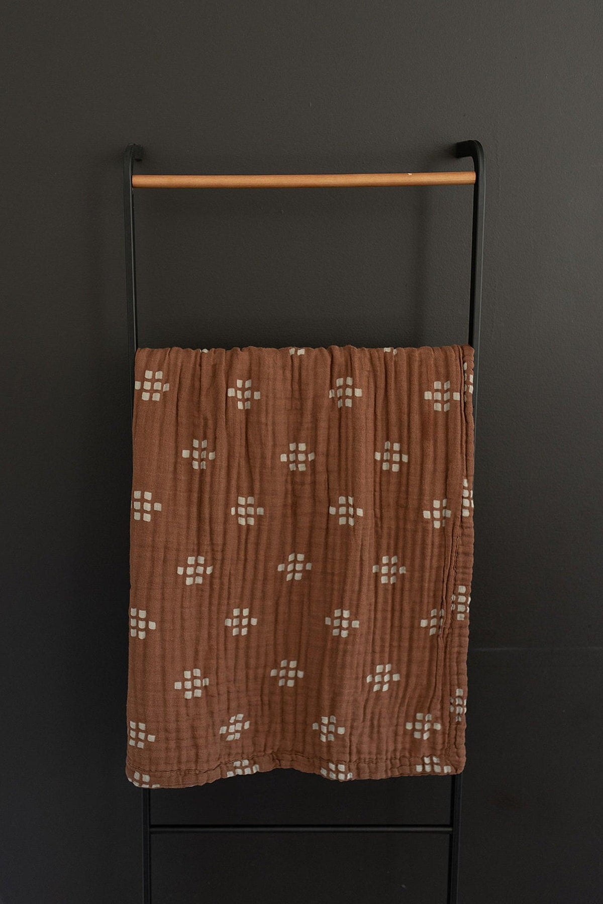 A brown muslin quilt with white geometric patterns hangs on a black metal rack against a dark wall.