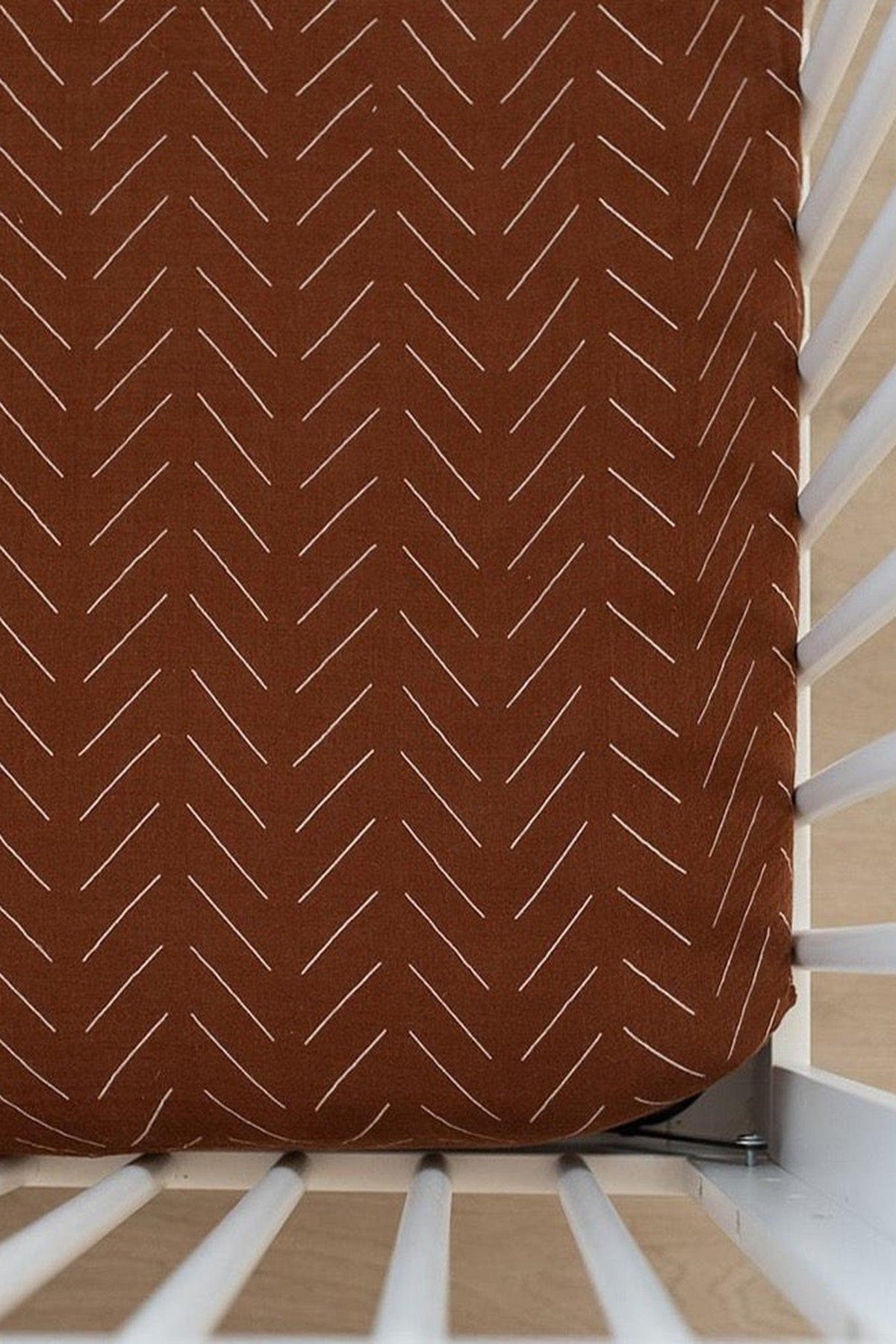Rust-colored crib sheet with white chevron patterns in a white crib, showcasing a stylish and cozy nursery detail.