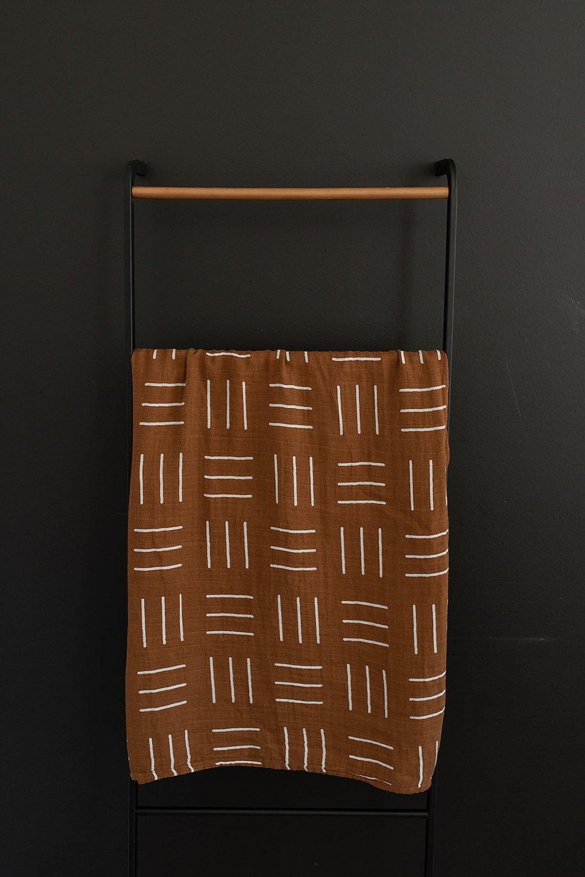 A mustard-colored muslin swaddle blanket with white geometric lines hangs on a black metal rack.