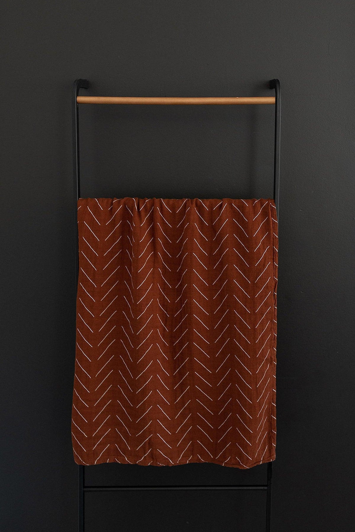 Rust-colored muslin swaddle blanket with white chevron patterns, hanging on a black and wooden rack.
