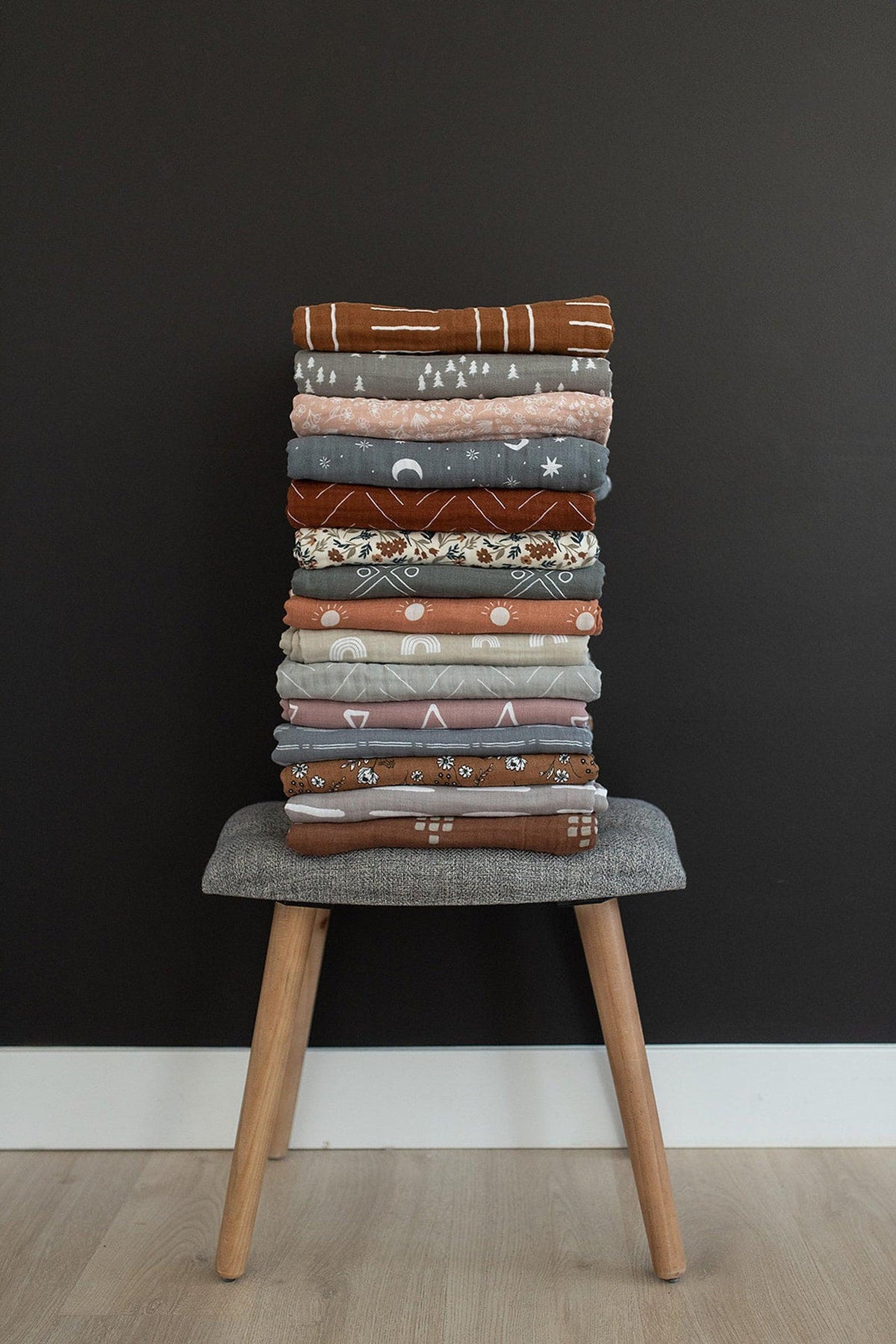 A stack of neatly folded patterned quilts in various earthy tones on a gray stool against a dark wall.