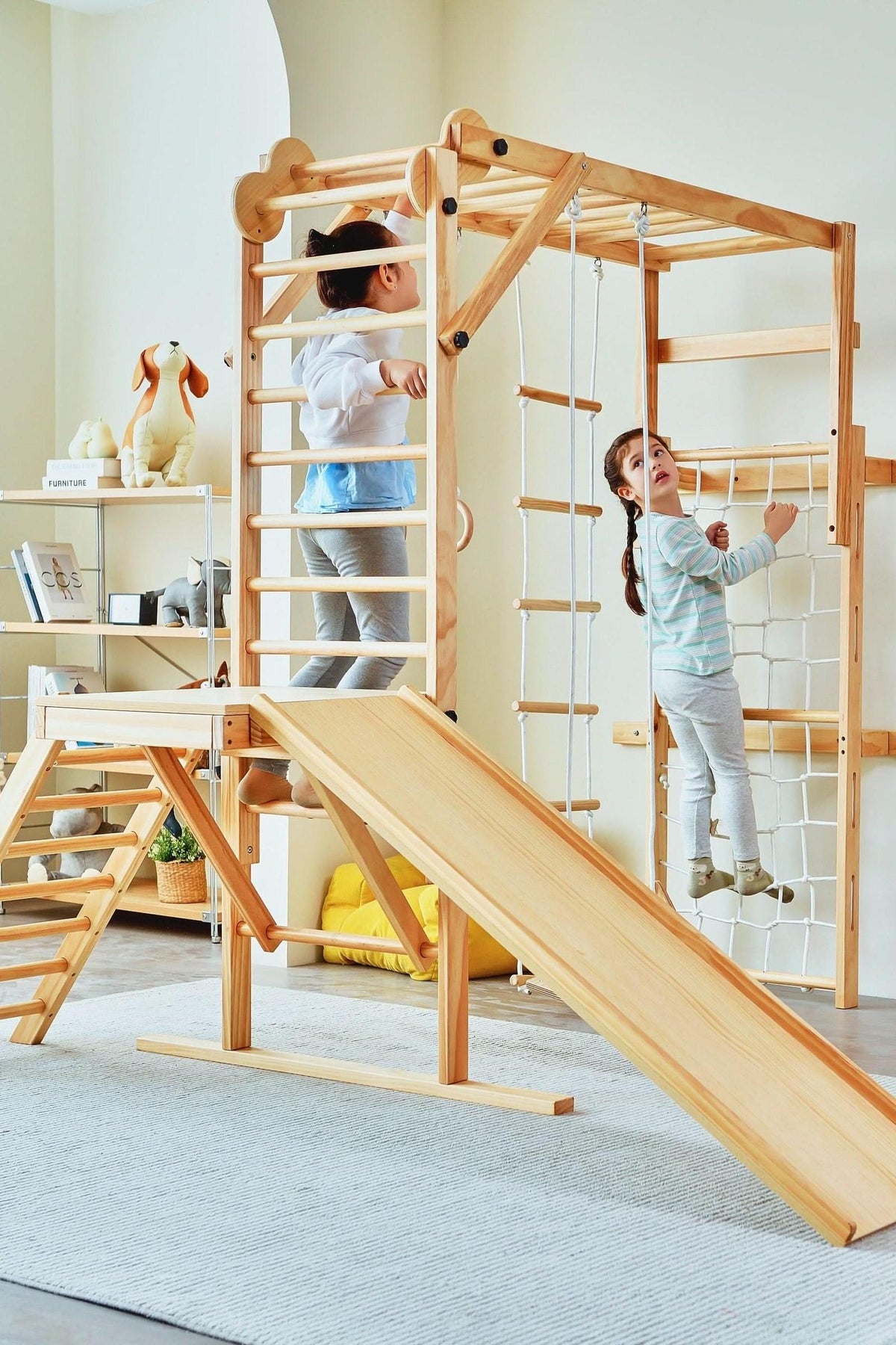 Two children play on a natural wood foldable jungle gym set featuring a slide, climbing ladder, and rope elements, set in a bright, airy indoor space.