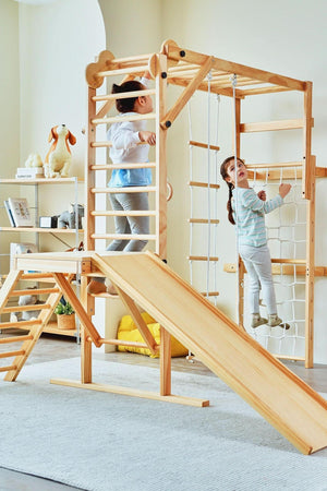 Two children play on a natural wood foldable jungle gym set featuring a slide, climbing ladder, and rope elements, set in a bright, airy indoor space.