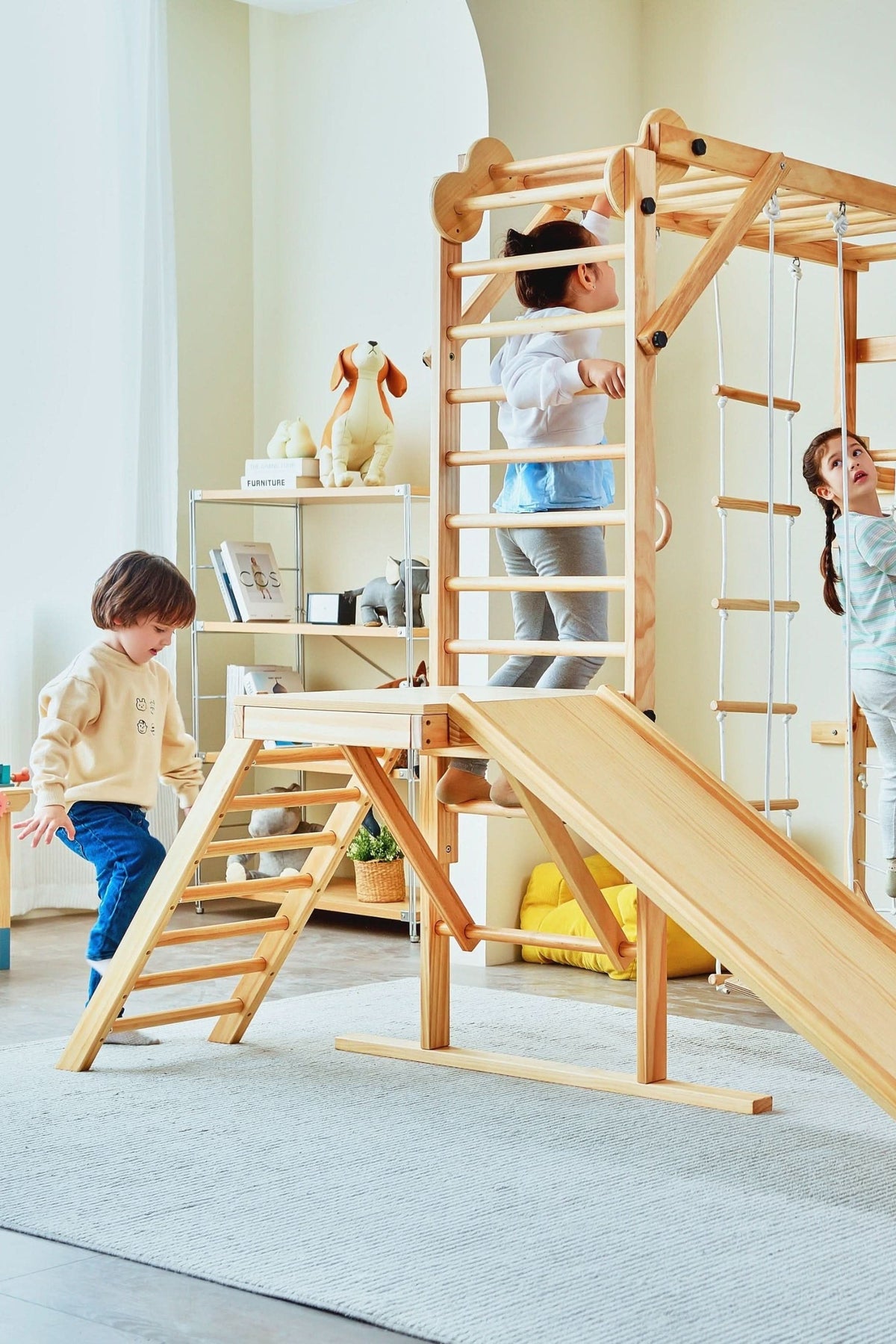 Two children play on a wooden indoor jungle gym with a slide, ladders, and climbing bars.