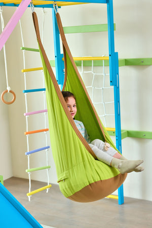 A child sits comfortably in a vibrant green hanging hammock chair, part of a colorful indoor playset. The playset features blue climbing bars, a rope net, and a swing.