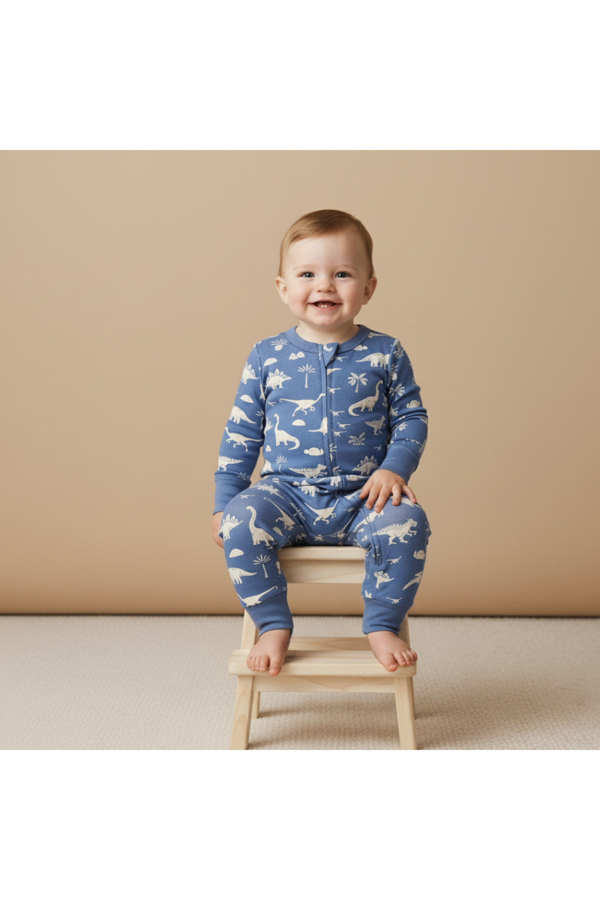 A smiling baby sits on a wooden stool, wearing a blue organic cotton romper with a playful dinosaur print. The soft fabric and two-way zipper ensure comfort and easy changing.