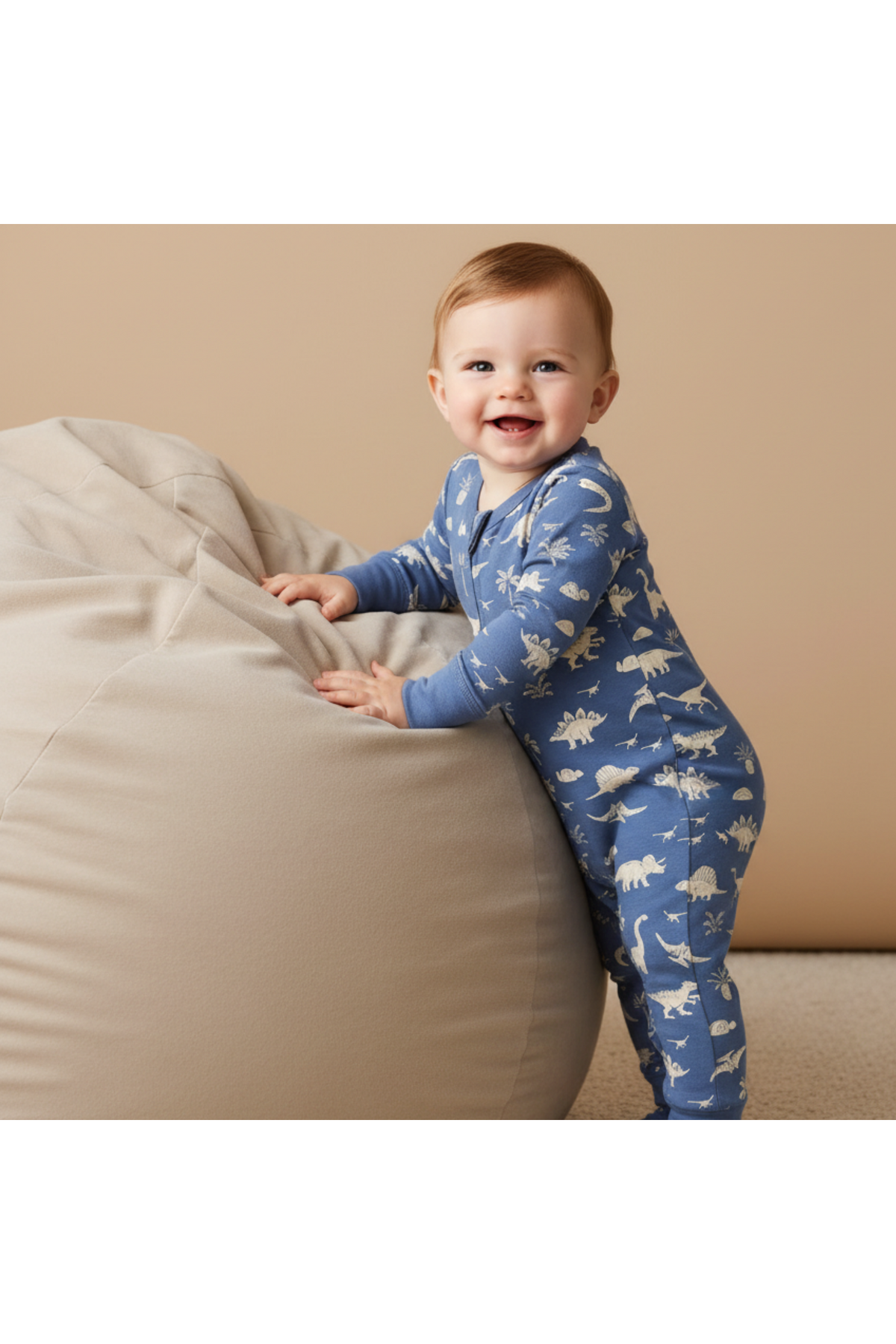 A smiling baby stands beside a large, soft beige bean bag, wearing a blue organic cotton romper featuring white dinosaur prints. The romper is designed for comfort and easy changing.