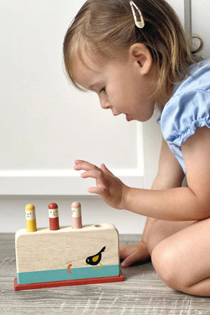A toddler interacts with a colorful wooden bird pop-up toy, featuring cheerful figures and a vibrant bird design. The eco-friendly toy encourages imaginative play and fine motor skills.