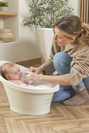 A woman gently bathes a baby in a Shnuggle Baby Bath, set on a wooden floor with plants nearby.