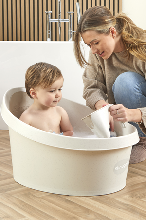 A toddler sits in a cream-colored Shnuggle bath tub while an adult pours water.