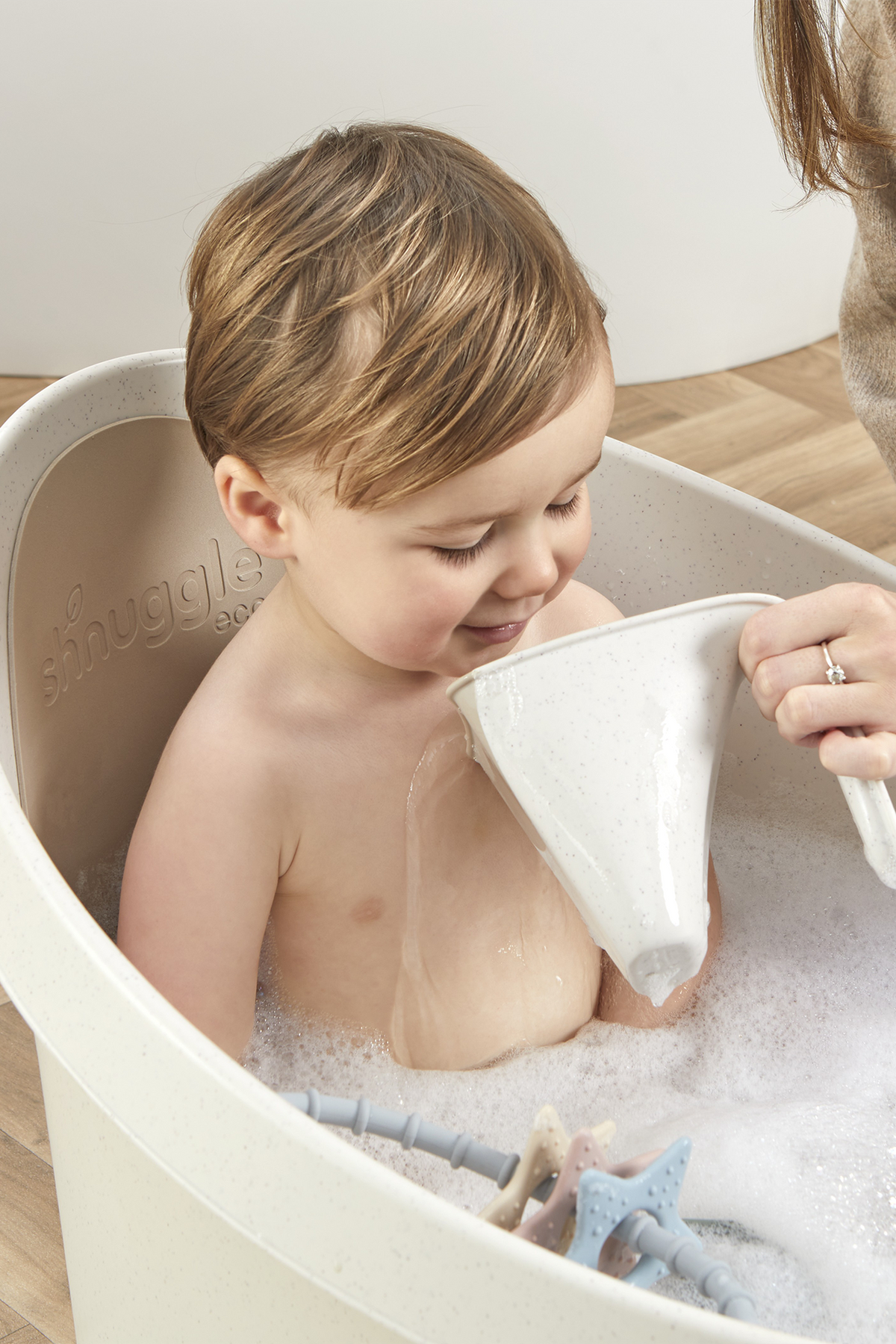 A toddler is enjoying a bath in a white Shnuggle tub, with water being poured gently over them.