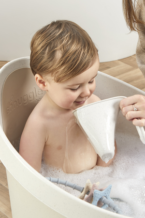 A toddler is enjoying a bath in a white Shnuggle tub, with water being poured gently over them.