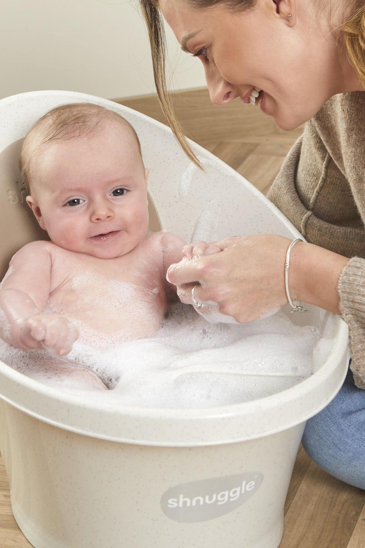 A baby sits in a Shnuggle bath filled with bubbles, smiling while a person gently washes their arm.
