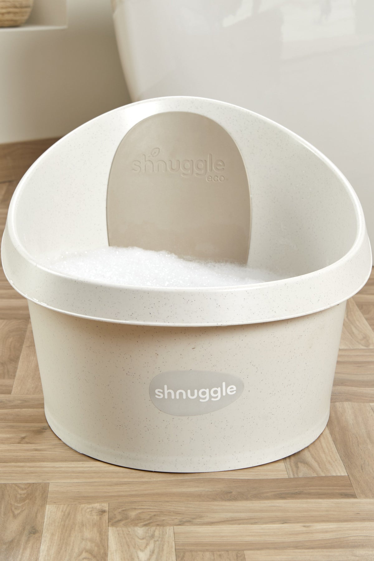 Cream-colored Shnuggle toddler bath tub with speckled texture and foam, set on a wooden floor.